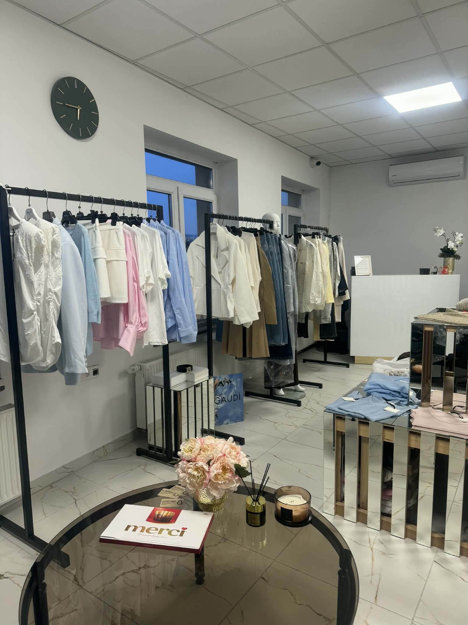 Wnętrze butiku MOORI — nowe kolekcje włoskiej odzieży na wieszakach, stolik z kwiatami i świecą zapachową